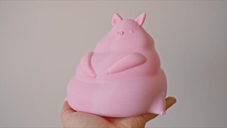 May include: A pink, 3D printed pig figurine with a rounded, plump body and small ears. The pig is sitting with its arms crossed in front of it.