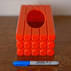 May include: Orange plastic tissue box cover with a rectangular opening and a textured bottom. The bottom is made of a series of small circles.