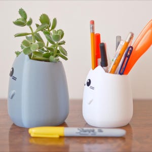 May include: Two cat-shaped containers, one light blue with a succulent plant and the other white with pens, pencils, and scissors. A yellow and gray Sharpie marker rests in front of them. The containers have black eyes and whiskers.