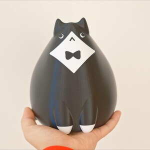 May include: A black cat figurine with a white bib and bow tie. The cat has white paws and ears. The figurine is held in a person's hand against a neutral background. The cat has a rounded body and a stylized design.