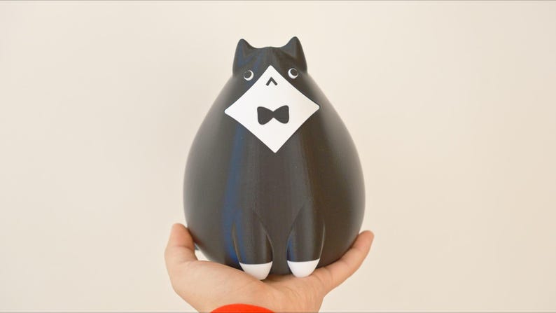 May include: A black cat figurine with a white bib and bow tie. The cat has white paws and ears. The figurine is held in a person's hand against a neutral background. The cat has a rounded body and a stylized design.