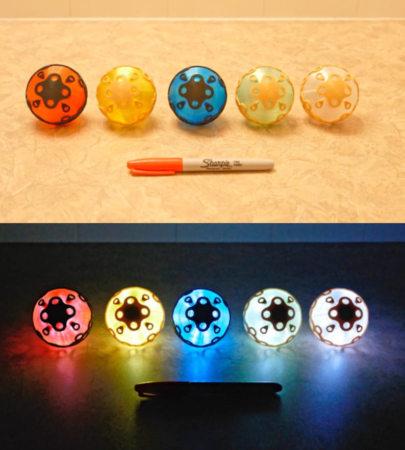 May include: A collection of five colorful, spherical toys with a patterned design, illuminated in various colors. An orange Sharpie marker is placed next to the toys. The bottom image shows the toys lit up with internal lights.