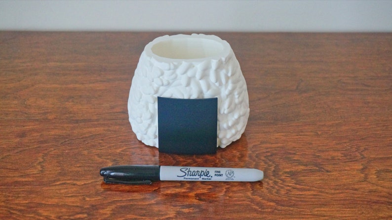 Puede incluir: Maceta de cer&aacute;mica blanca con forma de bola de arroz onigiri con una pizarra cuadrada negra en la parte delantera. Un marcador permanente negro Sharpie de punta fina se encuentra delante de la maceta.