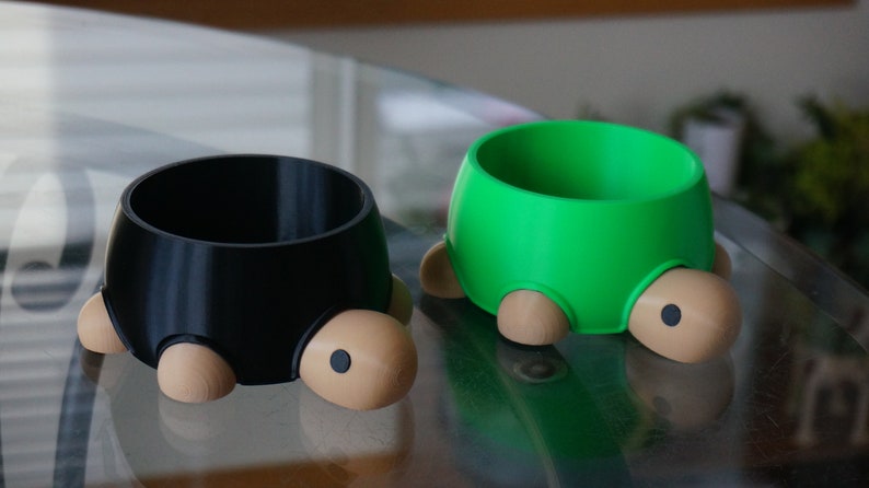 May include: Two turtle-shaped pet bowls, one black and one green, with brown legs and eyes. The bowls are made of plastic and are designed to look like turtles.
