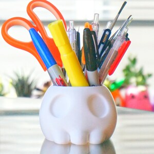 May include: A white ceramic skull-shaped pencil holder with a variety of pens, markers, and scissors sticking out of the top.