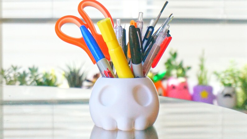 May include: A white ceramic skull-shaped pencil holder with a variety of pens, markers, and scissors sticking out of the top.