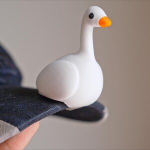 May include: A white goose figurine with an orange beak and black eyes sits on a dark blue baseball cap. The goose is perched on the brim of the cap, which has a gray and white trim.