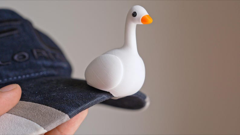 May include: A white goose figurine with an orange beak and black eyes sits on a dark blue baseball cap. The goose is perched on the brim of the cap, which has a gray and white trim.
