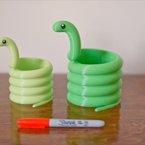 May include: Two green snake-shaped containers with coiled bodies and raised heads. One is a lighter shade of green than the other. A red and gray Sharpie marker is in front of the containers.