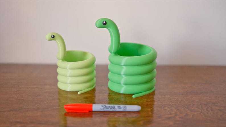 May include: Two green snake-shaped containers with coiled bodies and raised heads. One is a lighter shade of green than the other. A red and gray Sharpie marker is in front of the containers.