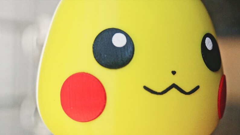 May include: Close-up of a yellow Pikachu figurine with black eyes, a red cheek, and a black smile.
