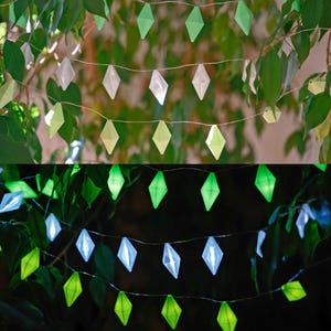 May include: Decorative string lights with diamond-shaped green and white accents. The lights are strung on a thin wire and are set against a backdrop of green leaves. The lights are illuminated in the bottom half of the image.