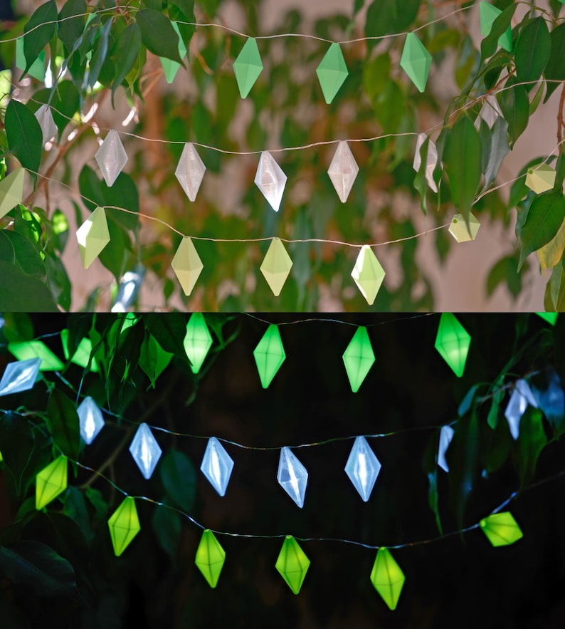 May include: Decorative string lights with diamond-shaped green and white accents. The lights are strung on a thin wire and are set against a backdrop of green leaves. The lights are illuminated in the bottom half of the image.