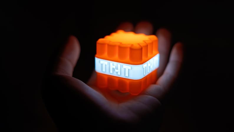 Op de afbeelding: Een kleine oranje kubus met een wit label waarop "TNT" staat. De kubus gloeit en wordt in de hand van een persoon gehouden.