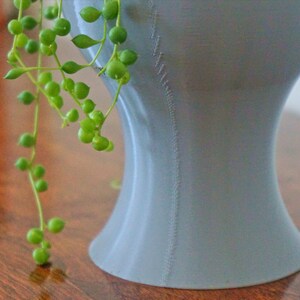 May include: A light gray ceramic planter with a unique, curved shape. The planter has a textured surface and is filled with a trailing green plant.