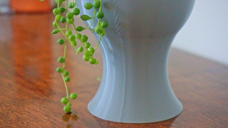May include: A light gray ceramic planter with a unique, curved shape. The planter has a textured surface and is filled with a trailing green plant.