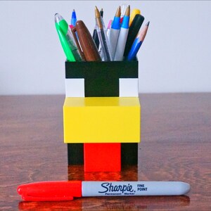 May include: A black, white, yellow, and red Lego pencil holder shaped like a Minecraft character. The holder is filled with pens and pencils.
