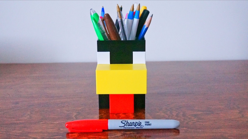 May include: A black, white, yellow, and red Lego pencil holder shaped like a Minecraft character. The holder is filled with pens and pencils.