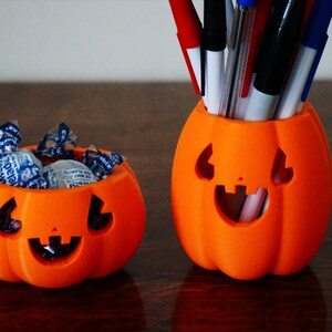 May include: Two orange pumpkin-shaped containers. The smaller container is filled with blue and white wrapped candies. The larger container is filled with pens and pencils.