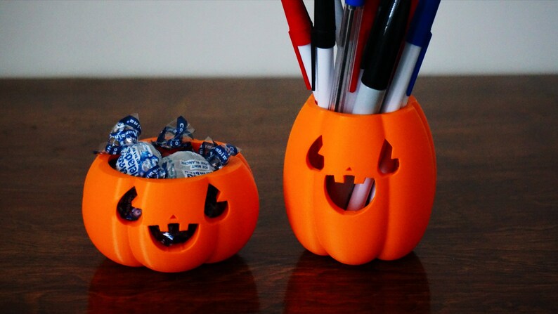 May include: Two orange pumpkin-shaped containers. The smaller container is filled with blue and white wrapped candies. The larger container is filled with pens and pencils.