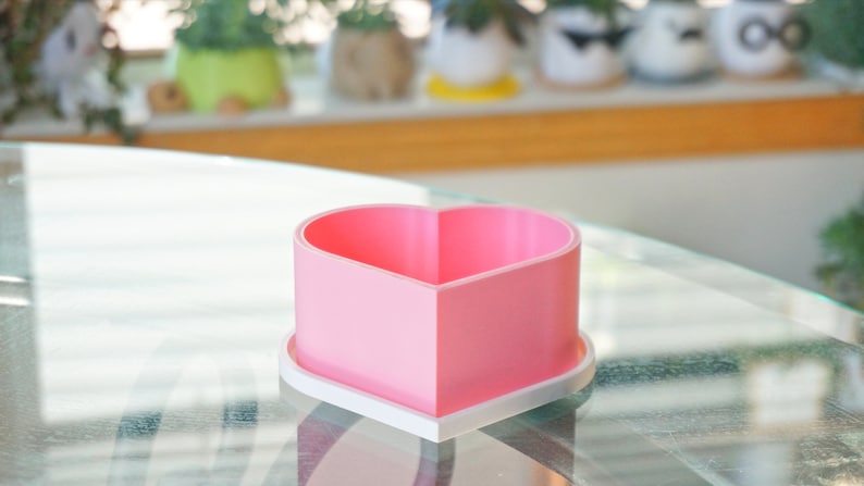 Puede incluir: Una maceta de coraz&oacute;n rosa con una base blanca. La maceta est&aacute; hecha de pl&aacute;stico y tiene un acabado liso.