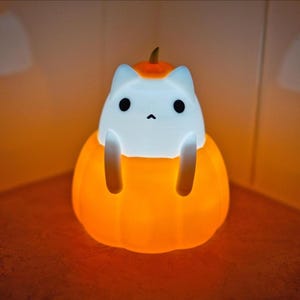 May include: A glowing orange and white cat-shaped night light. The cat figure sits inside a pumpkin, with the pumpkin and cat's face illuminated. The light is designed for Halloween or seasonal decor.