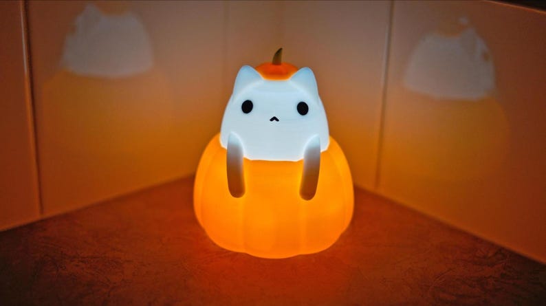 May include: A glowing orange and white cat-shaped night light. The cat figure sits inside a pumpkin, with the pumpkin and cat's face illuminated. The light is designed for Halloween or seasonal decor.