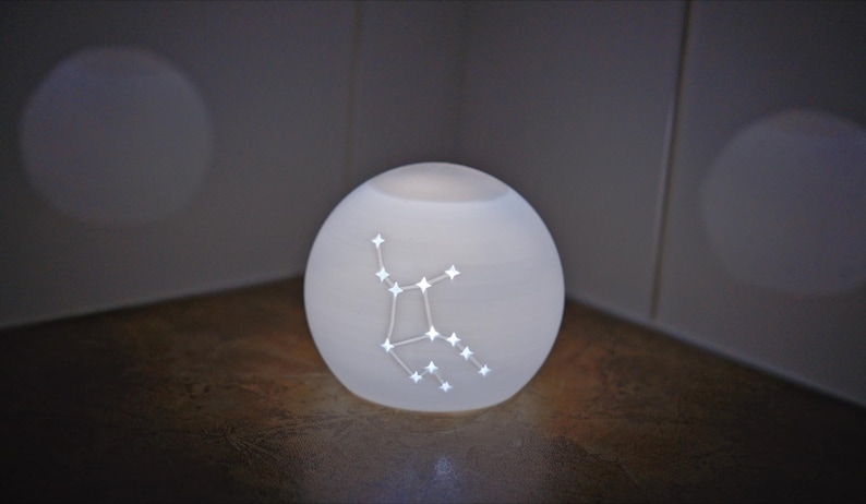 May include: A glowing white orb-shaped lamp with a constellation design. The constellation is illuminated with small, bright lights. The lamp sits on a wooden surface, and the background is a neutral color.