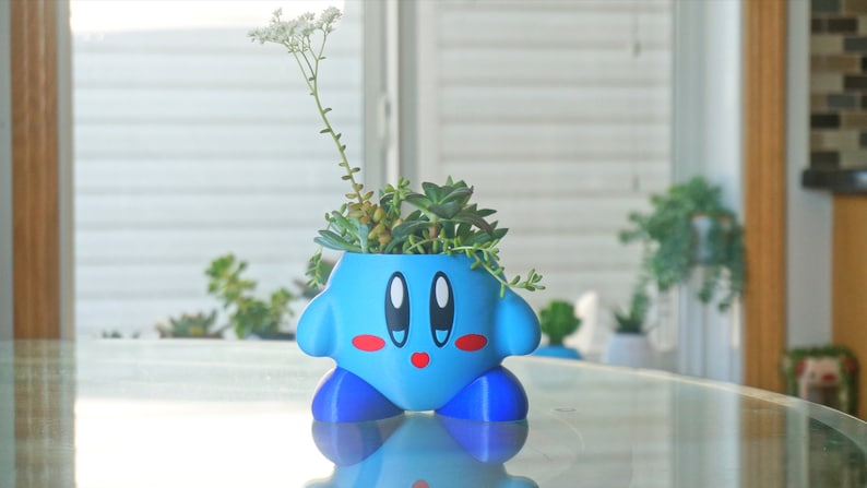 Puede incluir: Un macetero de personaje azul y rojo con una cara linda, que contiene una variedad de suculentas verdes y una planta con flores blancas.