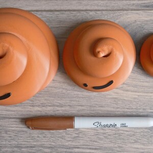 May include: Three brown ceramic poop emoji figurines of different sizes with black painted eyes and mouths. The figurines are arranged in a row on a wood surface with a white Sharpie fine point marker.