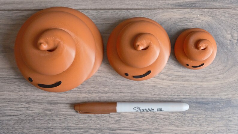 May include: Three brown ceramic poop emoji figurines of different sizes with black painted eyes and mouths. The figurines are arranged in a row on a wood surface with a white Sharpie fine point marker.