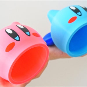 May include: Two pink and blue character-shaped cups with a large opening. The cups are made of plastic and feature a cartoon face with eyes, a mouth, and cheeks.