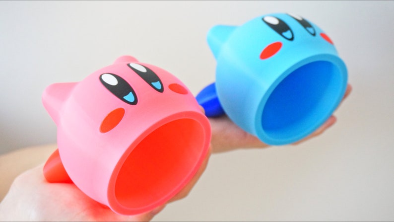 May include: Two pink and blue character-shaped cups with a large opening. The cups are made of plastic and feature a cartoon face with eyes, a mouth, and cheeks.