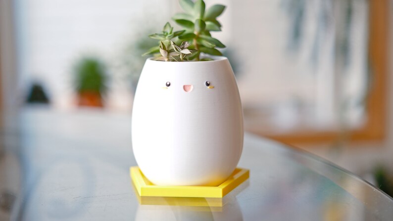 Op de afbeelding: Een witte keramische pot met een vrolijk gezicht en gele ogen, met een kleine groene succulent erin. De pot staat op een gele zeshoekige basis.