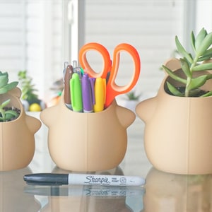 May include: Three beige ceramic planters shaped like cartoon animals, each with a different succulent plant. The middle planter has a set of colorful pens and orange scissors inside.