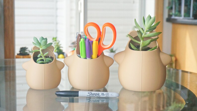 May include: Three beige ceramic planters shaped like cartoon animals, each with a different succulent plant. The middle planter has a set of colorful pens and orange scissors inside.