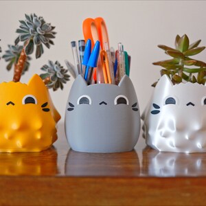 May include: Three cat-shaped ceramic planters, one yellow, one gray, and one white, with a succulent plant in each. The gray planter has a bunch of pens and pencils inside.