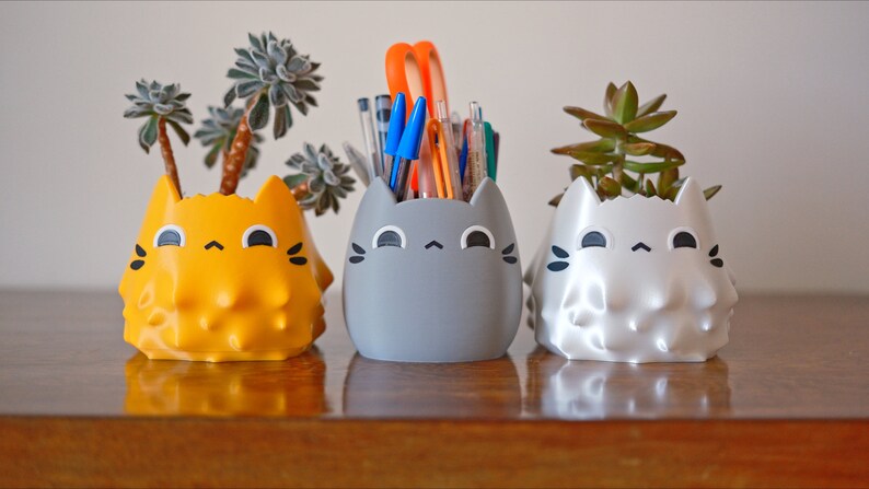 May include: Three cat-shaped ceramic planters, one yellow, one gray, and one white, with a succulent plant in each. The gray planter has a bunch of pens and pencils inside.