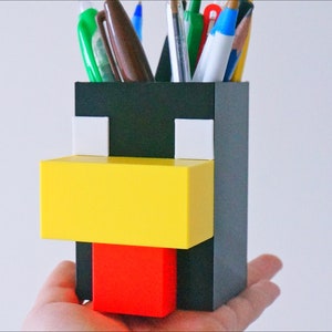 May include: A black, white, yellow, and red plastic pencil holder shaped like a Minecraft chicken.