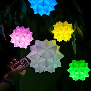 May include: Five colorful paper lanterns in the shape of a star, hanging from a tree branch. The lanterns are lit up and glow in different colors: blue, pink, yellow, white, and green. A remote control is in the foreground, suggesting the lanterns can be controlled.