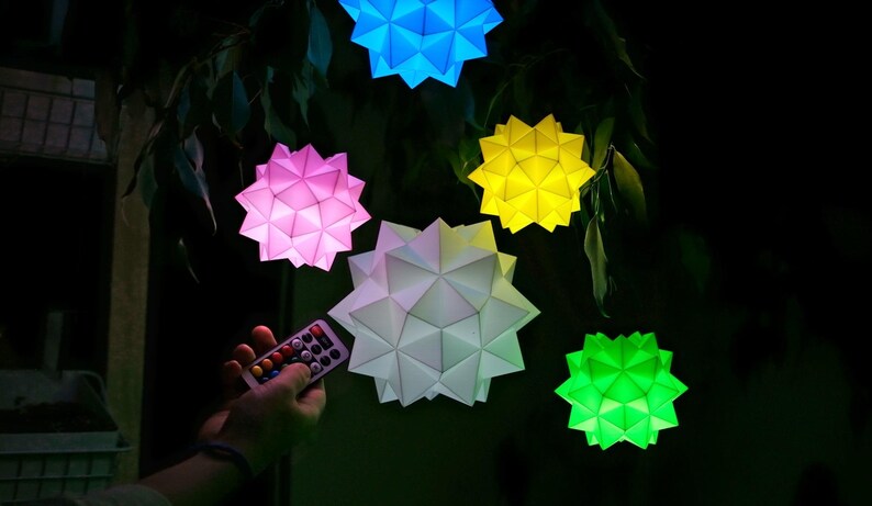 May include: Five colorful paper lanterns in the shape of a star, hanging from a tree branch. The lanterns are lit up and glow in different colors: blue, pink, yellow, white, and green. A remote control is in the foreground, suggesting the lanterns can be controlled.