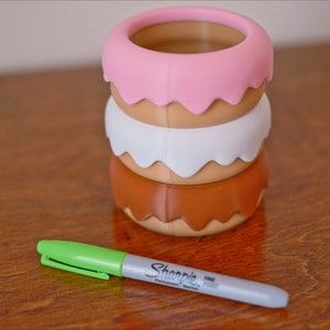 Puede incluir: Una pila de tres recipientes con forma de donut, cada uno con un glaseado de color diferente: rosa, blanco y marr&oacute;n. Los recipientes est&aacute;n hechos de un material liso, similar al pl&aacute;stico. Un marcador verde y gris est&aacute; sobre una superficie de madera.