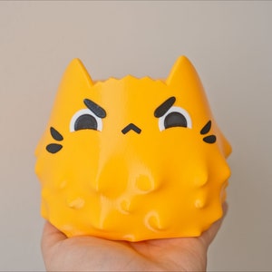 May include: A yellow ceramic planter shaped like a cat with a grumpy expression. The planter has black eyes, a black nose, and black whiskers.