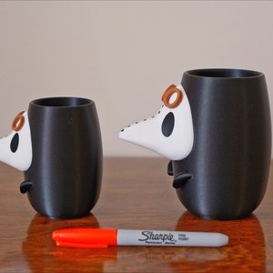 May include: Two black ceramic mugs with white and brown 3D animal faces. The mugs are different sizes and are sitting on a wooden surface with a red marker.