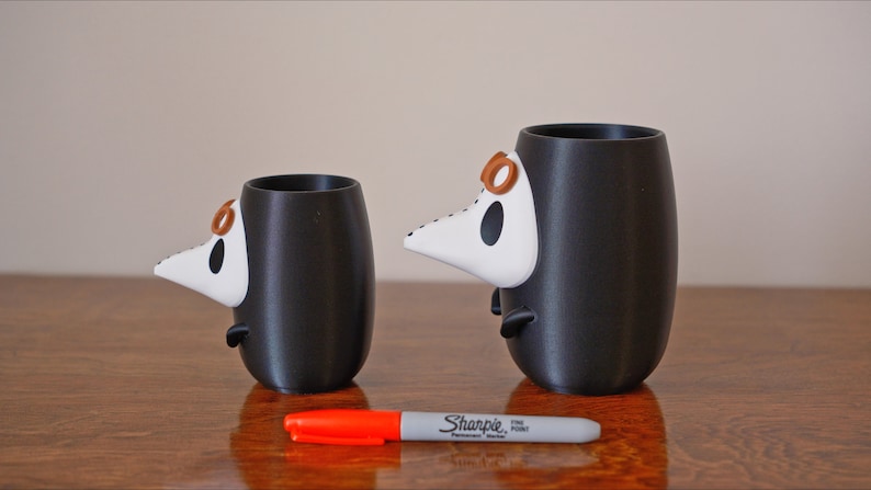 May include: Two black ceramic mugs with white and brown 3D animal faces. The mugs are different sizes and are sitting on a wooden surface with a red marker.