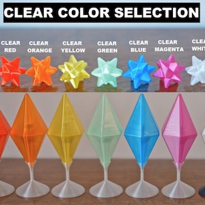 May include: A selection of eight clear plastic diamond-shaped ornaments in various colors, including red, orange, yellow, green, blue, magenta, and white. The ornaments are displayed on a white surface with a black background.