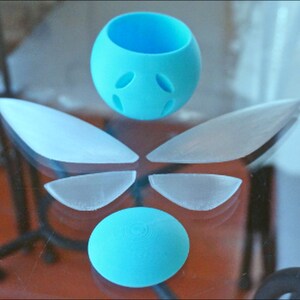 Puede incluir: Un conjunto de cinco piezas de pl&aacute;stico azul claro, que incluyen dos alas grandes, dos alas m&aacute;s peque&ntilde;as y una esfera con agujeros. Las piezas est&aacute;n dispuestas en un patr&oacute;n sim&eacute;trico sobre una superficie de vidrio.