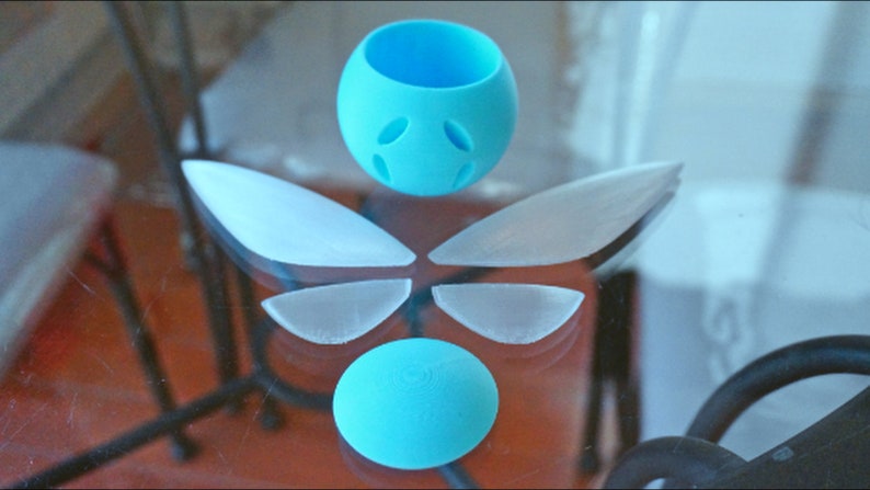 Puede incluir: Un conjunto de cinco piezas de pl&aacute;stico azul claro, que incluyen dos alas grandes, dos alas m&aacute;s peque&ntilde;as y una esfera con agujeros. Las piezas est&aacute;n dispuestas en un patr&oacute;n sim&eacute;trico sobre una superficie de vidrio.