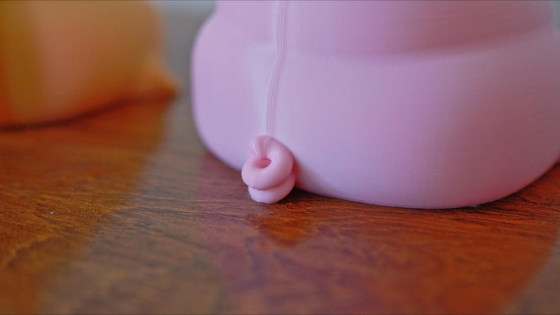 May include: A pink pig figurine with a curled tail. The pig is made of a smooth, soft material and has a rounded shape.