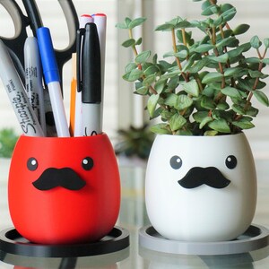 May include: Two white ceramic planters with black mustaches and googly eyes. One planter is red and holds pens and scissors. The other planter is white and holds a small green plant.